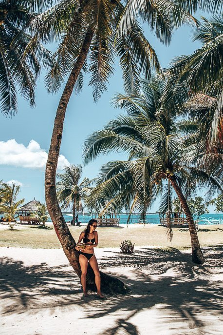 A girl in a black bikini holds a coconut standing in the shade of palm trees on Daku Island