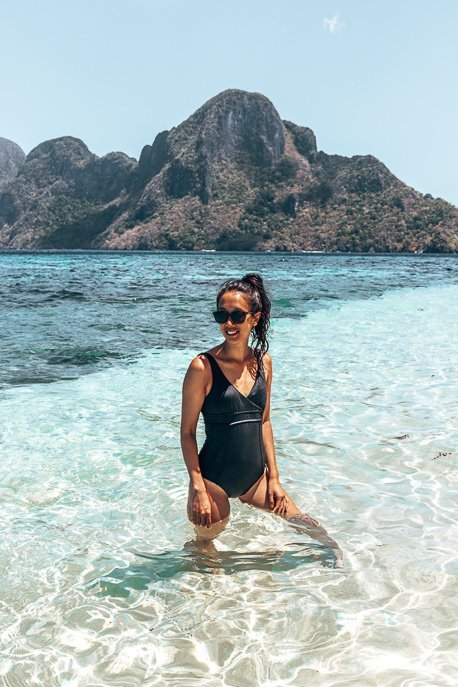 A girl in a black one piece swimsuit and sunglasses smiles while posing in the shallow waters of El Nido