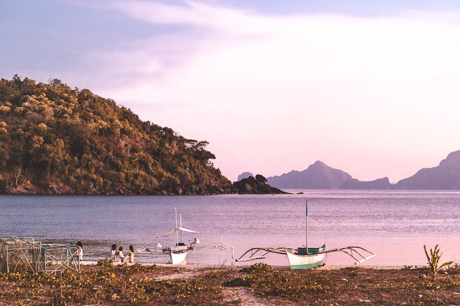 Young girls playing by the water next to two boats at Nacpan Beach during sunset, El Nido