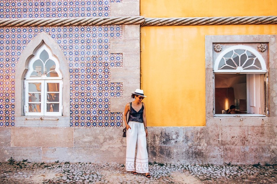 A woman poses in front of the yellow and blue walls of Pena Palace in Sintra, Portugal
