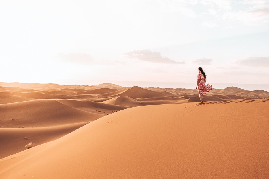 A woman in a red dress stands on a sand dune looking out into the distance in the Sahara Desert, Morocco