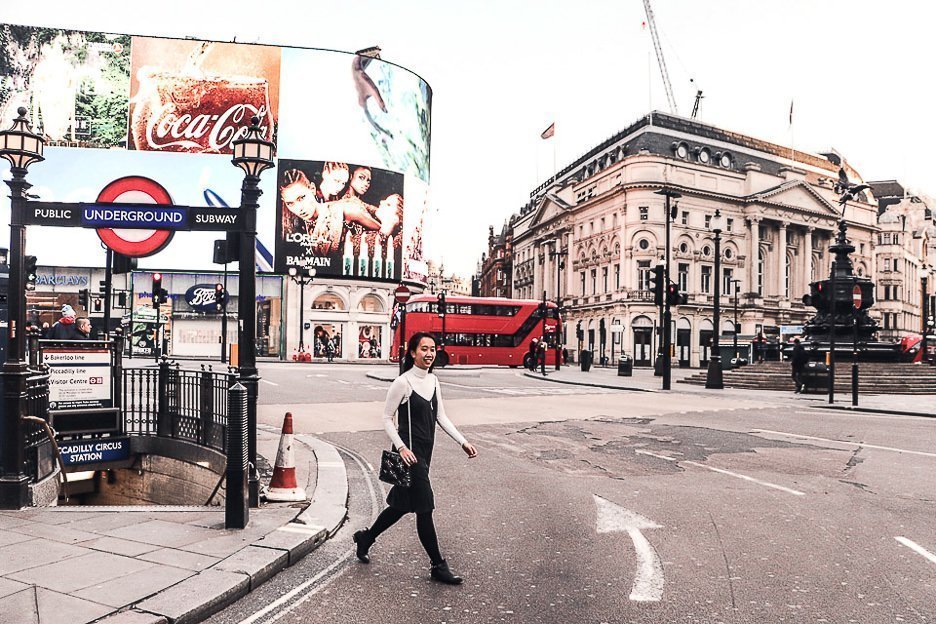 Jasmine of The Travel Quandary walks through Piccadilly Circus on a winter's day. The TV screens and a red double decker bus is in the background. London - United Kingdom