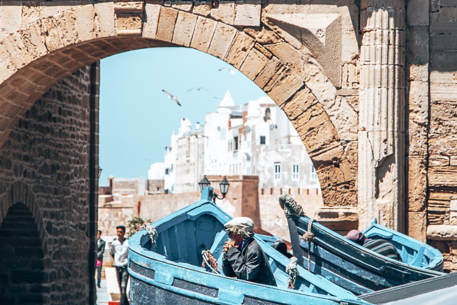 An old Moroccan man sits inside a blue boat ready to pose for photos for tourists at Essaouira harbour, Morocco