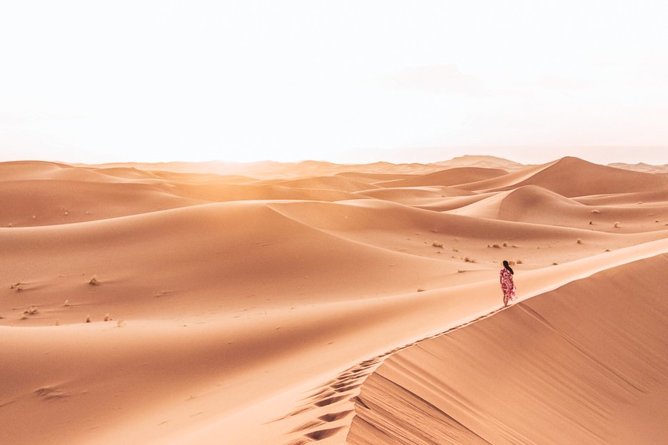 A woman in a red dress walks on the edge of a sand dune in the Sahara Desert, Morocco