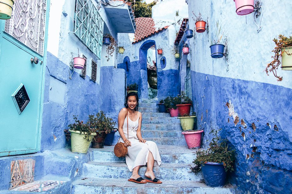 Jasmine of The Travel Quandary sits on the steps of a lane with coloured potted plants in Chefchaouen Morocco