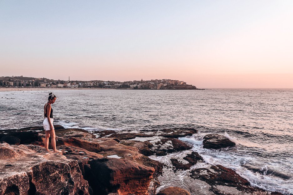 A girl stands on the rocks of Bondi Beach with a peachy sunrise on the horizon, Bondi Beach, New South Wales, Australia