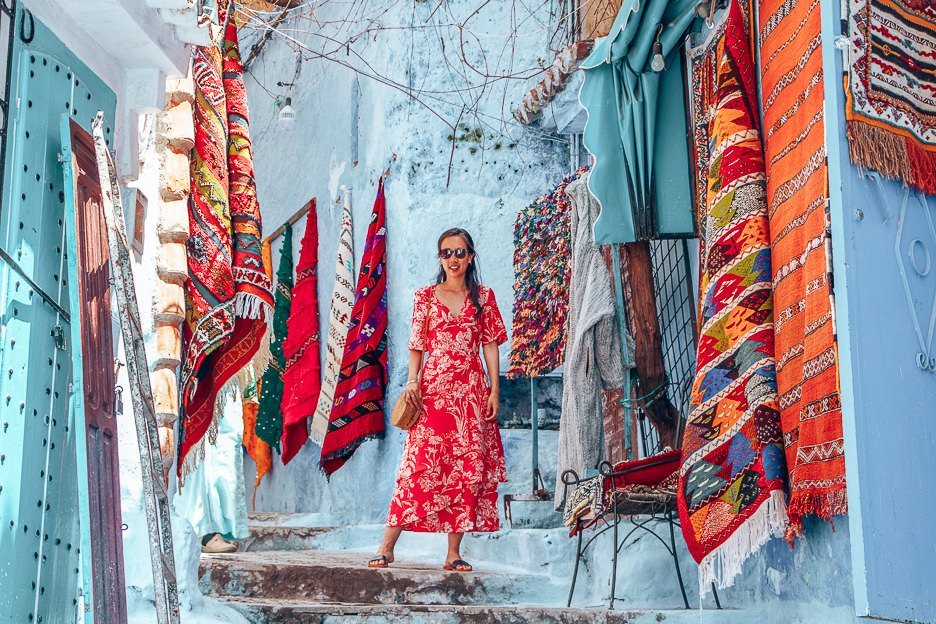 Jasmine of The Travel Quandary poses in front of woven Moroccan rugs for sale, Chefchaouen Morocco