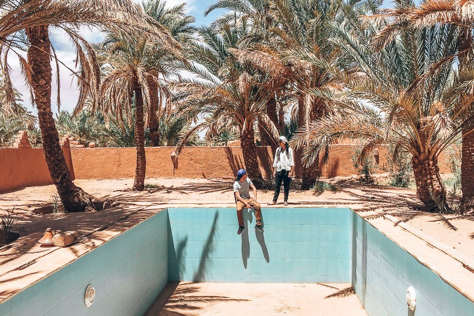 A man and a woman don Berber scarves and stand by an empty pool with palm trees in the Sahara Desert, Morocco