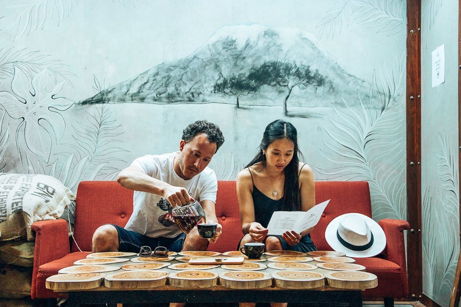 A man pours coffee while a woman reads the menu. Sitting on a red couch in Fabrica Coffee Roasters in Lisbon, Portugal